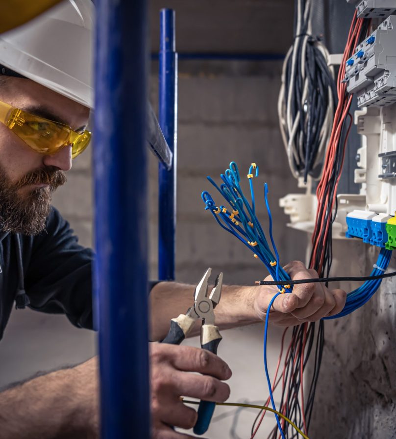 A male electrician works in a switchboard with an electrical connecting cable, connects the equipment with tools.
